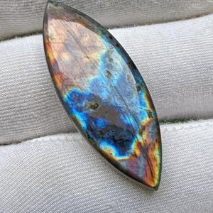 May include: A marquise-shaped labradorite gemstone featuring a polished surface with iridescent flashes of electric blue, gold, and orange hues. The stone displays natural internal inclusions and a smooth, reflective finish across its elongated form.