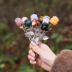 May include: A bouquet of gemstone roses in various colours, including pink, green, red, and black. The roses are mounted on silver stems with leaves. The bouquet is held in a person's hand.