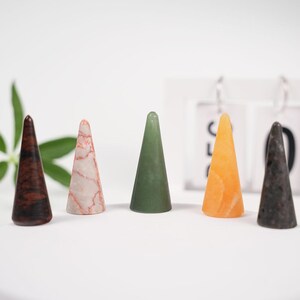 Può includere: Cinque coni di pietre preziose lucide in vari colori: marrone scuro, rosa e bianco, verde, arancione e grigio scuro. I coni sono disposti in fila su una superficie bianca. Le pietre hanno una finitura liscia e lucida.