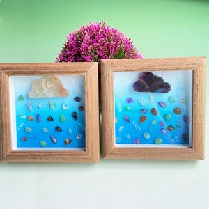 May include: Two framed art pieces with wooden frames. Each features a cloud-shaped stone at the top and colorful stones resembling raindrops against a blue gradient background. A cluster of purple flowers sits behind the frames.