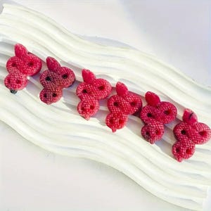 May include: Six red snake-shaped hair clips on a white, wavy surface. Each clip features a textured, scale-like pattern and a small, rounded head. The clips are a vibrant shade of red.