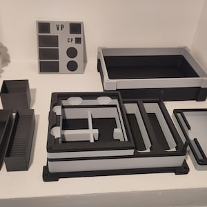 Puede incluir: Un surtido de contenedores y organizadores de almacenamiento grises y negros. La colección incluye bandejas rectangulares, cajas y un panel con las letras "VP" y "CP". Los artículos están dispuestos sobre una superficie blanca.