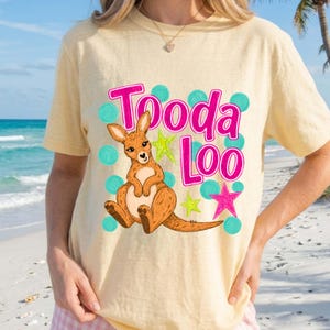 Può includere: T-shirt giallo chiaro con la scritta rosa "Tooda Loo" e un'illustrazione di un canguro. Il design include pois turchese e stelle verdi e rosa. La maglietta è in stile casual.