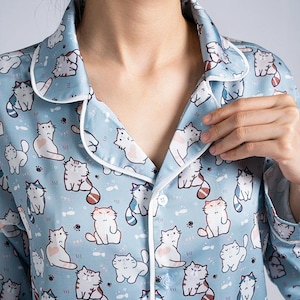 Op de afbeelding: Lichtblauw pyjamajasje met een herhaald patroon van cartoonkatten. De top heeft witte randen rond de kraag en een knoopsluiting. De stof lijkt een zacht, zijdeachtig materiaal te zijn.