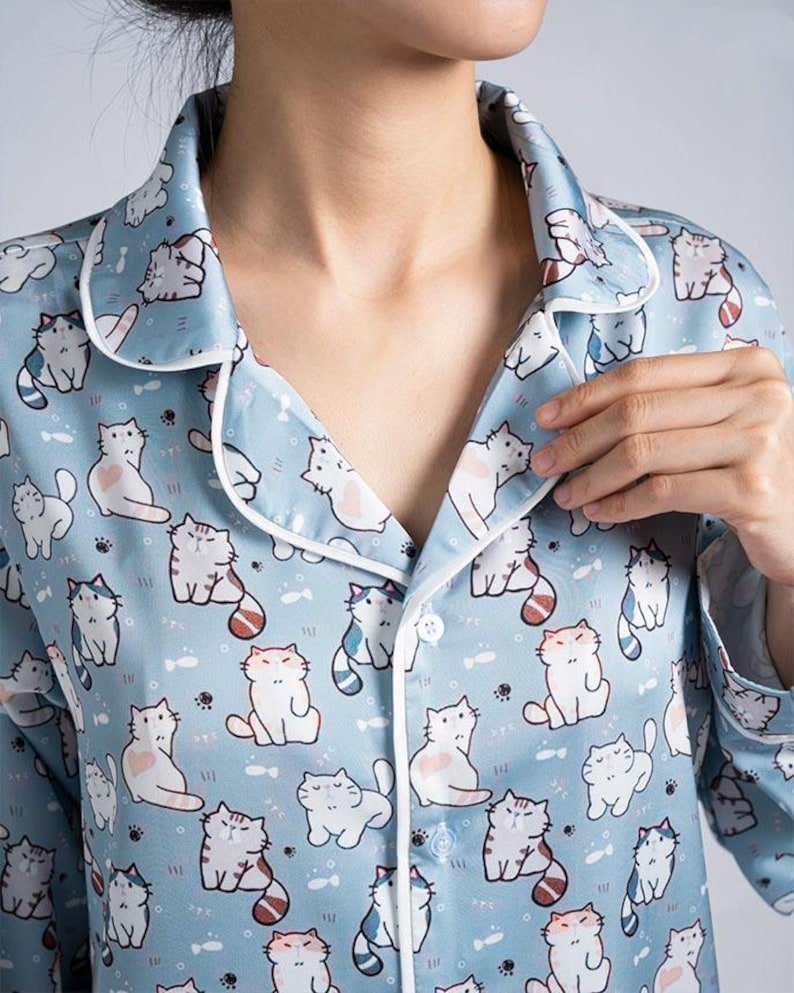 Op de afbeelding: Lichtblauw pyjamajasje met een herhaald patroon van cartoonkatten. De top heeft witte randen rond de kraag en een knoopsluiting. De stof lijkt een zacht, zijdeachtig materiaal te zijn.