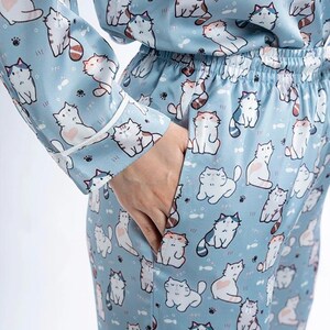 Op de afbeelding: Lichtblauwe pyjamaset met een herhalend patroon van cartoonkatten in verschillende poses. De top met lange mouwen en de broek hebben witte biezen en een taille met trekkoord. De katten zijn wit, grijs en oranje, met hart- en visdetails.