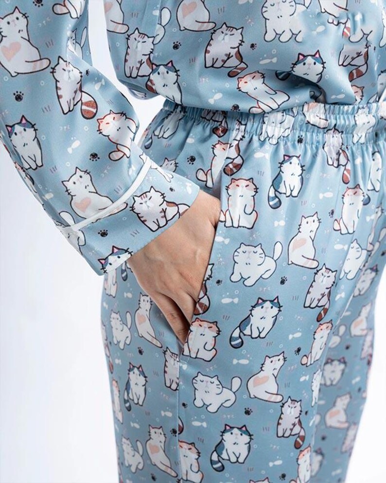 Op de afbeelding: Lichtblauwe pyjamaset met een herhalend patroon van cartoonkatten in verschillende poses. De top met lange mouwen en de broek hebben witte biezen en een taille met trekkoord. De katten zijn wit, grijs en oranje, met hart- en visdetails.