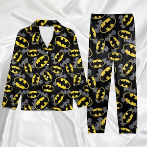 May include: Black pajama set with a repeating pattern of yellow Batman logos and grey bat silhouettes. The top is a button-down shirt with a white collar. The pajama trousers have an elasticated waistband. The set is made of a soft material.