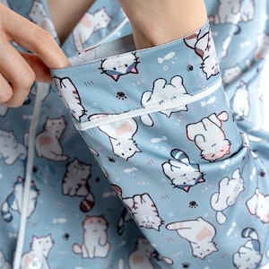 Op de afbeelding: Lichtblauwe pyjamatop met een herhaald patroon van witte cartoonkatten. De stof heeft een satijnglans en de manchetten en de bies zijn wit. De katten hebben roze accenten en verschillende poses.