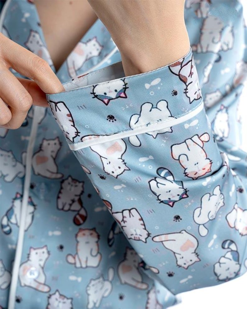 Op de afbeelding: Lichtblauwe pyjamatop met een herhaald patroon van witte cartoonkatten. De stof heeft een satijnglans en de manchetten en de bies zijn wit. De katten hebben roze accenten en verschillende poses.
