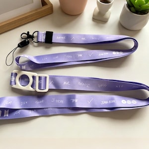 Op de afbeelding: Drie lavendelkleurige lanyards met witte tekst en accenten. Eén lanyard heeft een zwarte clip en een zwart touwtje. De lanyards hebben witte tekst met namen en symbolen. De lanyards zijn gemaakt van glad satijn.