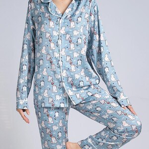 Op de afbeelding: Lichtblauwe pyjamaset met een herhaald patroon van witte en grijze cartoonkatten. De top met lange mouwen en de broek hebben witte biezen. De pyjama heeft een top met knopen en een kraag.