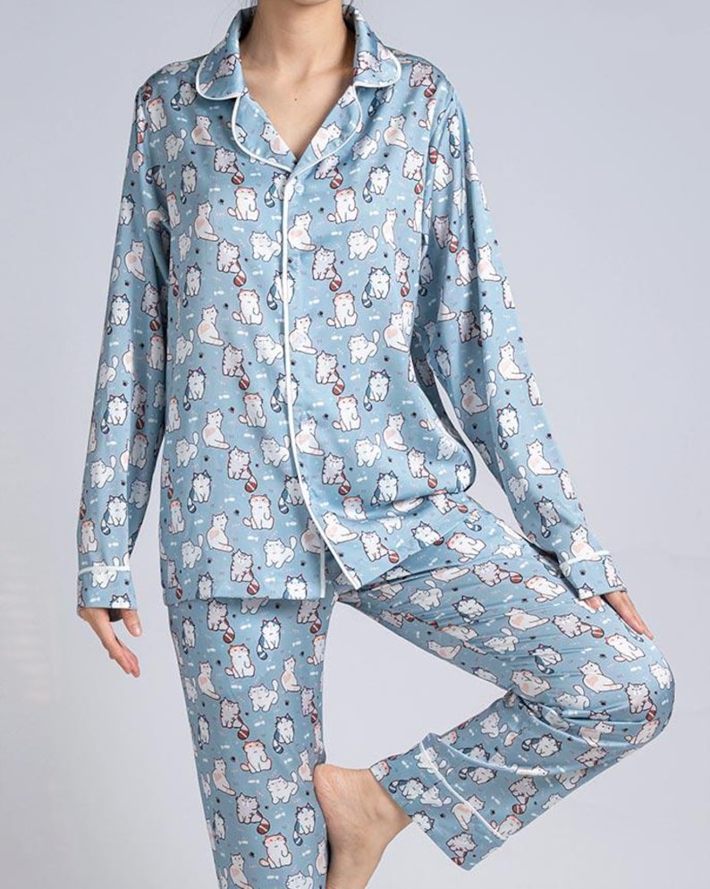 Op de afbeelding: Lichtblauwe pyjamaset met een herhaald patroon van witte en grijze cartoonkatten. De top met lange mouwen en de broek hebben witte biezen. De pyjama heeft een top met knopen en een kraag.
