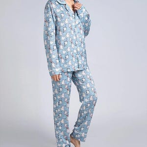 Op de afbeelding: Lichtblauwe pyjamaset met een herhaald patroon van witte kattenillustraties. De top met lange mouwen heeft een knoopsluiting en een reverskraag. De broek is lang en recht. Witte biezen accentueren de kraag, manchetten en broekzomen.