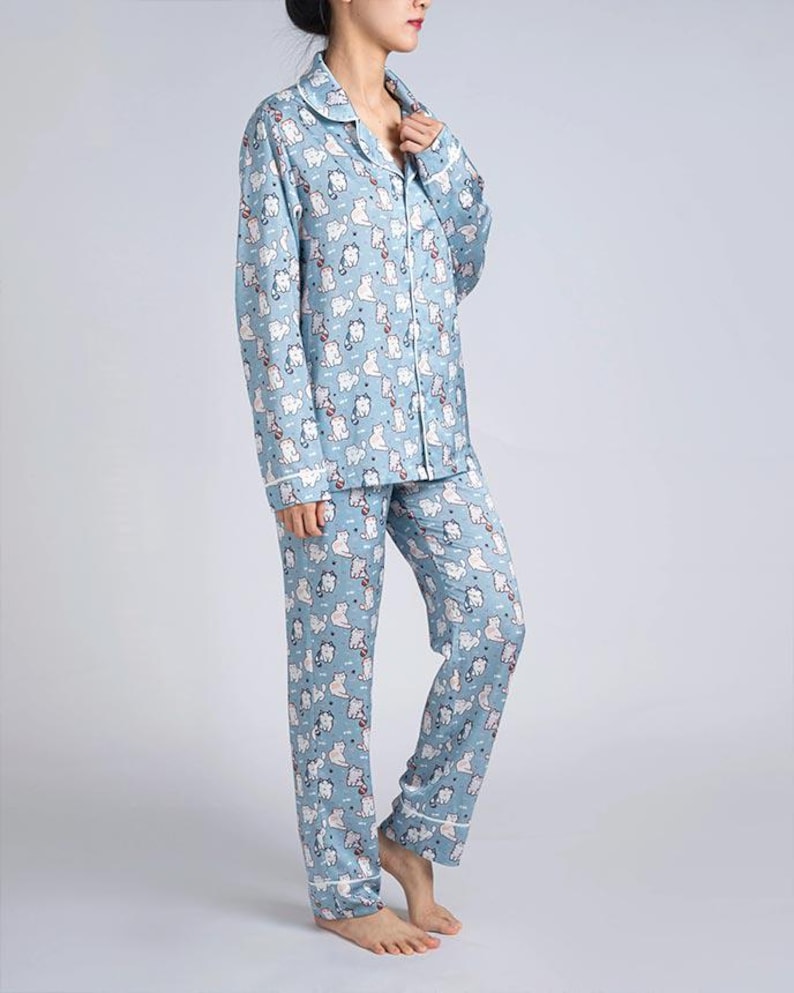 Op de afbeelding: Lichtblauwe pyjamaset met een herhaald patroon van witte kattenillustraties. De top met lange mouwen heeft een knoopsluiting en een reverskraag. De broek is lang en recht. Witte biezen accentueren de kraag, manchetten en broekzomen.