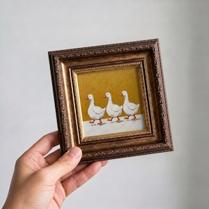 Op de afbeelding: Een ingelijst schilderij toont drie witte ganzen met oranje snavels en poten, lopend in een rij. Het kunstwerk staat tegen een gouden achtergrond en een witte voorgrond, omlijst door een sierlijke bruine lijst.