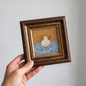 Op de afbeelding: Een ingelijst olieverfschilderij toont een witte eend met oranje voeten die in blauw water duikt. Het kunstwerk is gevat in een decoratieve, sierlijke, bruin en goudkleurige lijst. Het schilderij heeft een textuur, impasto stijl.