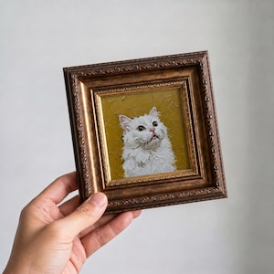 Könnte beinhalten: Gerahmtes Ölgemälde einer weißen Katze mit flauschigem Fell, vor einem senfgelben Hintergrund. Die Katze blickt nach oben, der Mund leicht geöffnet. Der Rahmen ist verziert, mit einem dunkelbraunen, goldfarbenen Finish.