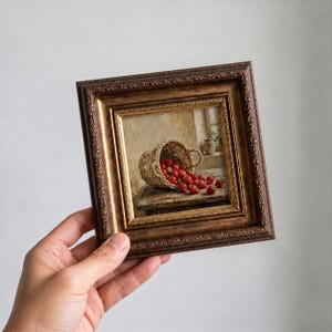 May include: A framed painting shows a woven basket spilling red strawberries. The painting is set in a rustic scene with a wooden table and a window in the background. The frame has an ornate gold and brown finish.