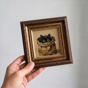 May include: A framed painting depicts a black cat peeking out of a brown paper bag. The cat's yellow eyes are visible. The painting is in a decorative, ornate, brown and gold frame.