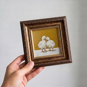 Puede incluir: Pintura enmarcada con dos patitos blancos. La obra de arte tiene un fondo dorado y una base blanca, con los patitos de pie juntos. El marco es marrón oscuro y ornamentado.