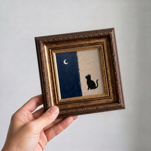 May include: A framed painting depicting a black cat against a beige backdrop, with a dark blue night sky and a crescent moon. The artwork is presented in a decorative gold-toned frame.