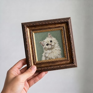 May include: A framed painting of a fluffy white cat with green eyes. The cat is set against a muted green background. The frame is ornate with a dark brown finish. The artwork is held by a hand.