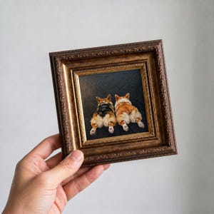Op de afbeelding: Een ingelijst schilderij met twee corgi-honden van achteren, tegen een donkere achtergrond. Het kunstwerk wordt gepresenteerd in een sierlijke, goudkleurige lijst, die een vleugje elegantie aan het stuk toevoegt.