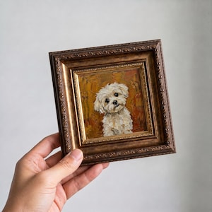 May include: A small square oil painting featuring a white fluffy dog with dark eyes and a nose, set against a textured golden-brown background. The artwork is encased in an ornate, dark bronze-colored frame with intricate embossed detailing.
