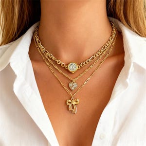 May include: A layered gold necklace set featuring a watch pendant, a heart pendant with small clear stones, and a bow pendant. The necklaces are displayed against a white shirt.