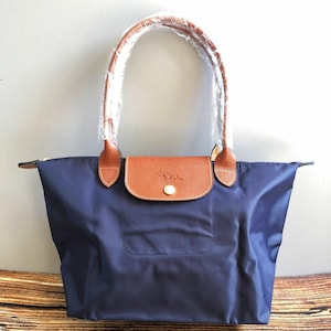 May include: Navy blue tote bag with brown leather accents. The bag features a brown leather flap with a gold-colored button closure. The handles are brown leather and wrapped in clear plastic. The bag is made of a smooth, durable material.
