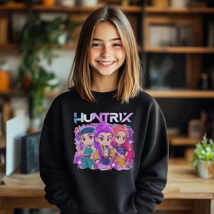 May include: A black sweatshirt featuring the word "HUNTRIX" in a blue and purple font. Below the text is a graphic of three cartoon characters with colorful hair and outfits. The characters are surrounded by stars and a purple background.