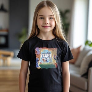May include: A young person wearing a black t-shirt with the text "YEEPS Hide & Seek" and a colorful cartoon graphic. The graphic features various cartoon characters in a playful scene. The person is smiling and has long blonde hair.