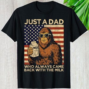 May include: Black t-shirt featuring a Bigfoot graphic wearing sunglasses and holding a milk carton. The design includes an American flag background and the text "JUST A DAD WHO ALWAYS CAME BACK WITH THE MILK."