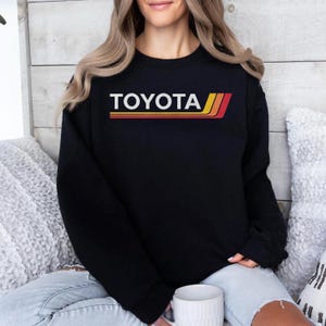 May include: Black crewneck sweatshirt with the word "TOYOTA" in white block letters. Below the text are three horizontal stripes in red, yellow, and orange. The sweatshirt is worn by a person sitting on a couch.