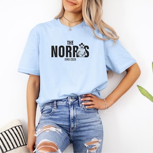 May include: Light blue t-shirt with the text "The Norris" in black block letters, and a graphic of a cowboy. The text "1940-2026" is below the name. The shirt is made of a soft material.