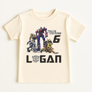 May include: Cream-colored short-sleeved t-shirt featuring a graphic of Transformers characters. The design includes the text "TRANSFORMERS 6" and the name "LOGAN" in black block letters. The Transformers logo is also present.