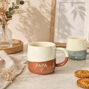 May include: Two ceramic mugs. One is cream and brown with "PAPA" and a heart. The other is cream and blue-green with a name and heart. Both have a curved design and a handle. The mugs are on a table.