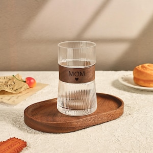 May include: A clear glass with vertical ridges, a brown leather band with the word "MOM" and a heart, sits on a wooden oval tray. The glass is partially filled with a clear liquid. A pastry is on a white plate.
