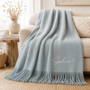 May include: A light blue throw blanket featuring the name Sophia embroidered in white cursive script followed by a small white heart outline. The blanket has a smooth texture and is finished with long, matching fringe along the bottom edge.