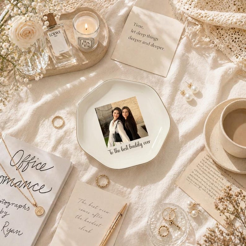 Puede incluir: Una composici&oacute;n plana con un plato de cer&aacute;mica blanco con una foto y la inscripci&oacute;n "To the best buddy ever". Otros elementos incluyen una vela, perfume, joyas, un libro y caf&eacute;, todo dispuesto sobre una superficie color crema.