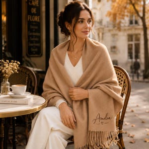 May include: A tan-coloured, soft-textured shawl featuring a fringe trim at the bottom edge. The fabric is embroidered with the name Amelia in a cursive script, accompanied by a small heart symbol centred below the name.