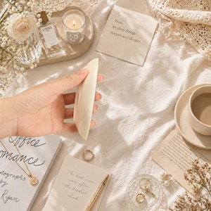 Puede incluir: Un objeto alargado de color crema se sostiene en una mano, rodeado de una colecci&oacute;n de art&iacute;culos. Se ven un frasco de perfume, una vela encendida y una taza de caf&eacute;. El texto "Time, let deep things deeper and deeper" est&aacute; en una tarjeta.