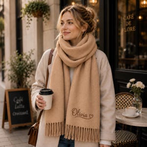 May include: A thick, tan-coloured scarf featuring a soft, brushed texture and long fringe detailing at the ends. The scarf is embroidered with the name Olivia in a cursive script, accompanied by a small heart symbol near the bottom edge.