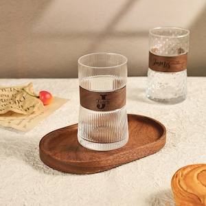 May include: Clear glass tumblers with wooden bands engraved with names and initials, displayed on a wooden tray. One tumbler has vertical ridges, the other has a textured pattern. The tray and bands are a warm brown color.