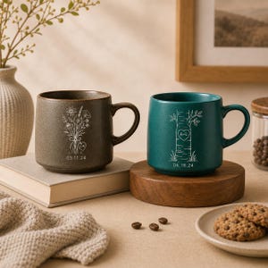 Puede incluir: Dos tazas de cerámica con asas. La taza izquierda es marrón con un dibujo lineal blanco de un ramo de flores silvestres, un corazón con las letras E+N y la fecha 05.11.24. La taza derecha es verde azulado con un árbol, un corazón con A+L y 06.18.24.