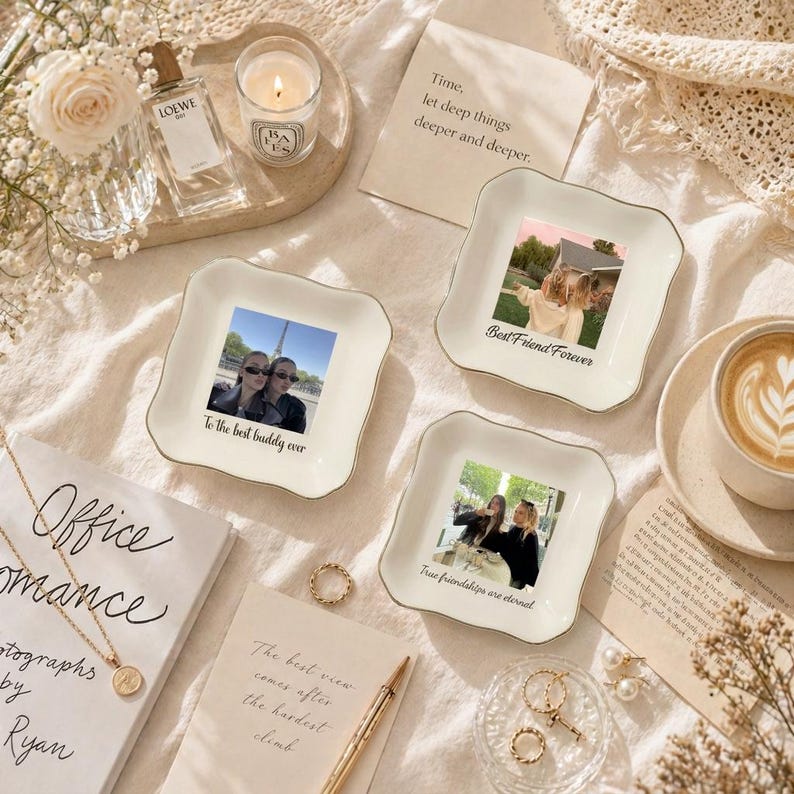 Puede incluir: Una colecci&oacute;n de platos decorativos de cer&aacute;mica blanca con ribete dorado, cada uno con una fotograf&iacute;a y una inscripci&oacute;n sobre la amistad. La escena incluye una vela, perfume, joyas y un libro.