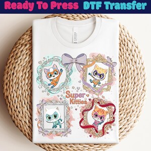 Peut inclure: T-shirt blanc avec un motif de quatre chats de dessin animé dans des cadres ornés, avec le texte "Super Kitties". Le motif comprend des nœuds et des étoiles. Le t-shirt est présenté sur une surface tissée.