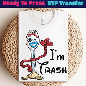 May include: White t-shirt featuring a cartoon character with a fork-like head, red arms, and the text "I'm Trash" in black. The shirt is folded and placed on a woven, circular surface.