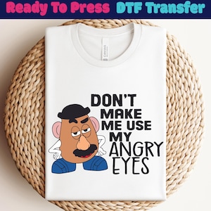 May include: White t-shirt with a graphic of a cartoon potato head character and the text "DON'T MAKE ME USE MY ANGRY EYES". The shirt is folded and placed on a woven, circular surface.